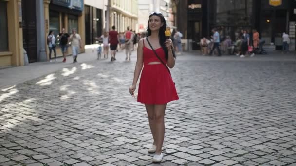 在阳光明媚的夏日午后 身穿红衣的年轻漂亮女子在布拉迪斯拉发历史城区的街道上散步时 在慢镜头下笑着吃着冰淇淋 — 图库视频影像