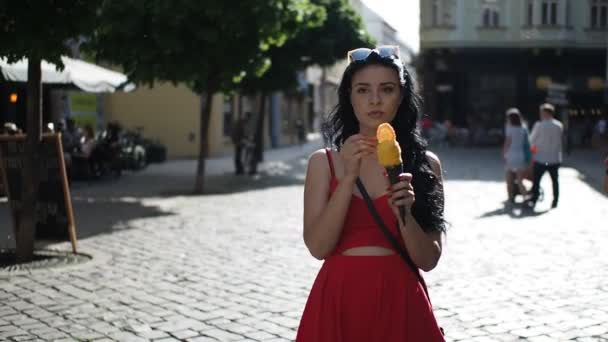 赤いドレスの若い美容女性は スローモーションHdビデオで晴れた夏の午後にブラチスラバ市の歴史的な部分の通りを歩いている間に笑顔とアイスクリームを食べています — ストック動画