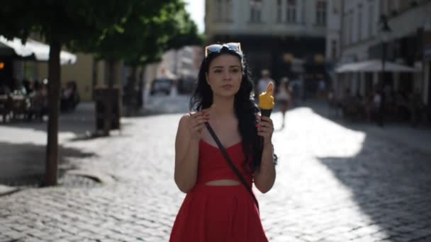 Mladá krasavice v červených šatech se usmívá a jí zmrzlinu při procházce po ulici v historické části Bratislavy za slunečného letního odpoledne ve videu Slow Motion Hd. Poloviční rychlost. 