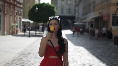 Kırmızı elbiseli genç bayan, güneşli bir yaz günü öğleden sonra Bratislava şehrinin tarihi bölgesinde yürürken gülümsüyor ve dondurma yiyor. Yarı hız. 