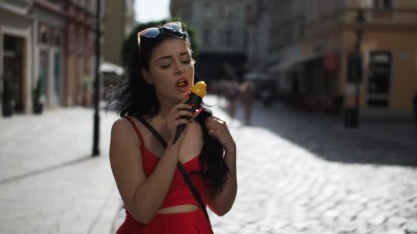 在阳光明媚的夏日午后 身穿红衣的年轻漂亮女子在布拉迪斯拉发历史城区的街道上观看慢镜头时 一边微笑着一边吃着冰淇淋 — 图库视频影像