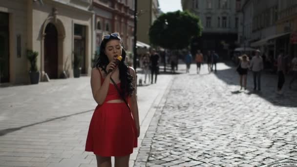 赤いドレスを着た若い美容女性は スローモーションHdビデオで晴れた夏の午後にブラチスラバ市の歴史的な部分の通りを見ている間に笑顔とアイスクリームを食べています — ストック動画