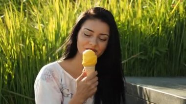 Genç güzel kadın Hd Video 'da dondurma yiyor. Sazlık yapraklarının ve nehir kıyısının önünde gülümsüyor ve sarı dondurmaları yalıyor. Düşük alan derinliği ve bulanık arkaplan.