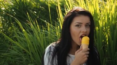 Genç güzel kadın Hd Video 'da dondurma yiyor. Sazlık yapraklarının ve nehir kıyısının önünde gülümsüyor ve sarı dondurmaları yalıyor. Düşük alan derinliği ve bulanık arkaplan.