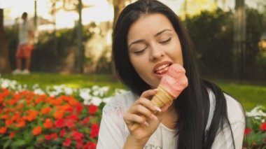 Genç güzel kadın Hd Video 'da dondurma yiyor. O, şehir parkında çiçek tarhının önünde gülümsüyor ve pembe dondurmaları yalıyor. Düşük alan derinliği ve bulanık arkaplan.