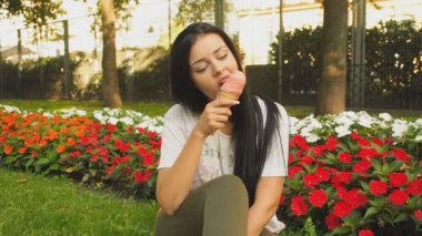 Genç güzel kadın Hd Video 'da dondurma yiyor. O, şehir parkında çiçek tarhının önünde gülümsüyor ve pembe dondurmaları yalıyor. Düşük alan derinliği ve bulanık arkaplan.