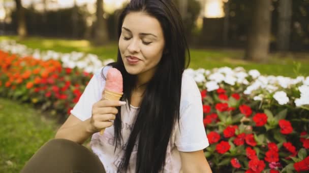 年轻的美女正在吃Hd视频中的冰淇淋 她笑着 在城市公园的花坛前舔着粉红色的明胶 实地深度低 背景模糊 — 图库视频影像