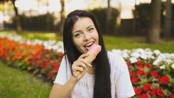 若い美人さんがHdビデオでアイスクリームを食べています 都会の公園の花壇の前でピンクのジェラートを笑って舐めています 被写界深度の低さと背景のぼやけ — ストック動画