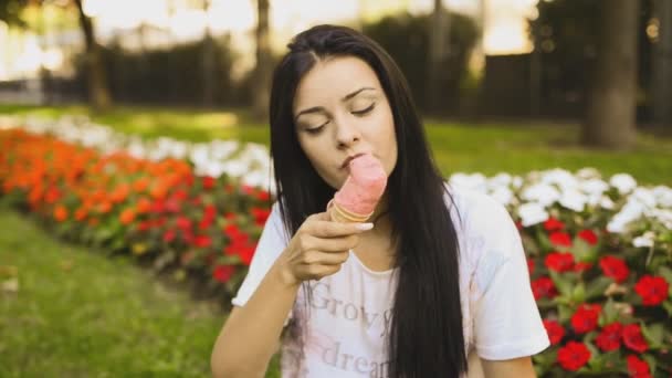 年轻的美女正在吃Hd视频中的冰淇淋 她笑着 在城市公园的花坛前舔着粉红色的明胶 实地深度低 背景模糊 — 图库视频影像