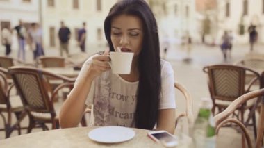 Genç güzel kadın HD VIDEO 'da terasta elinde kahveyle oturuyor. Kahve karıştırıyor, içiyor, akıllı telefona bakıyor ve gülümsüyor. Arka planda yürüyen insanlar..