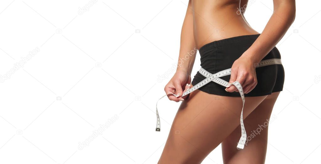 Weight loss - woman with slim body. Young skinny female in black shorts with tanned body is measuring her perfect buttocks with tape measure and showing diet result. Studio shot, white background.