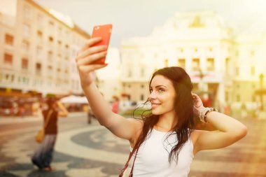 Gülümseyen ve selfie çeken güzel bir kız. Genç ve güzel bir kadın sıcak yaz öğleden sonra Bratislava şehrinin tarihi bölümünde kırmızı akıllı telefonuyla fotoğraf çekiyor..