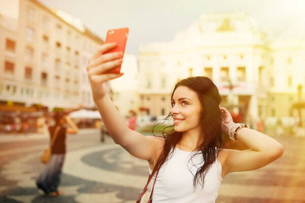 Красивая девушка улыбается и делает селфи. Молодая красивая женщина развлекается и фотографируется с красным смартфоном перед площадью в исторической части Братиславы в жаркий летний день
.
