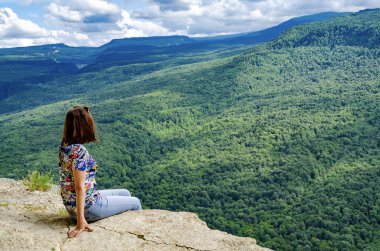 Genç kız uçurumun önündeki bir kayanın üzerinde oturuyor. Kolları güneşe doğru açık.