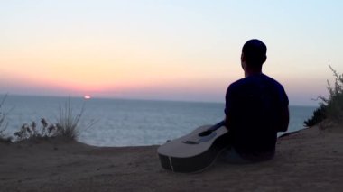 Gün batımında akustik gitar çalan genç bir adamın şarkı yazma silueti.