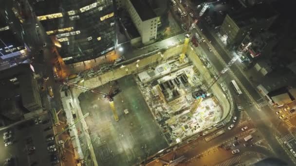 Escena nocturna en el sitio de construcción. Drone aéreo por encima de enormes construcciones con grúas y máquinas pesadas con paisaje urbano en el fondo — Vídeos de Stock