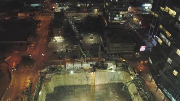 Scena notturna al cantiere. Drone aereo sopra enormi Costruzioni con gru e macchine pesanti con paesaggio urbano in background — Video Stock