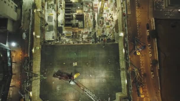 Ночная сцена на стройке. Воздушный беспилотник над огромными конструкциями с кранами и тяжелыми машинами с городским пейзажем на заднем плане — стоковое видео