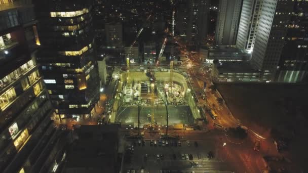 Scena nocna na budowie. Dron powietrzny nad ogromnymi konstrukcjami z dźwigami i ciężkimi maszynami z krajobrazem miejskim w tle — Wideo stockowe