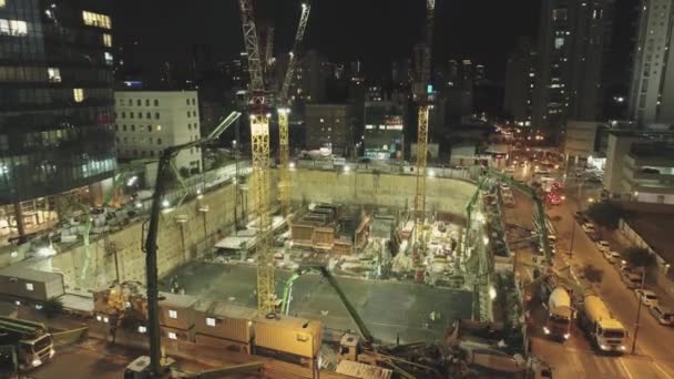 Cena noturna no canteiro de obras. Drone aéreo acima enorme Construções com guindastes e máquinas pesadas com paisagem urbana no fundo — Vídeo de Stock