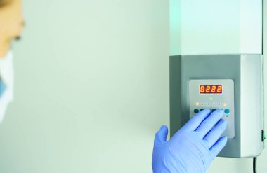 Sağlık görevlisi mikroplara karşı odayı dezenfekte etmek için lambayı yakar. Gösterge panelindeki düğmelere basar, mavi koruyucu bir eldiven verir.                 