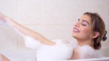 Güzel, kendine güvenen genç bir bayan köpükle banyo yapıyor, gülümsüyor ve gülüyor, vücuduyla ilgileniyor ve yıkıyor.