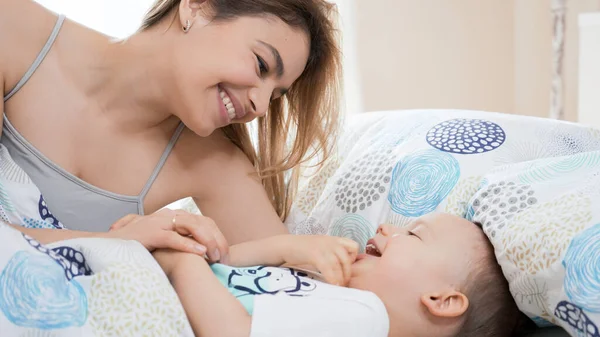 Güzel, genç bir anne ve küçük bir çocuk sabah birlikte eğleniyorlar. Gülümsüyor, gülüyor ve çocukla yatakta oynuyor. Aile mutluluğu konsepti. Parlak, güneşli sıcak bir gün..