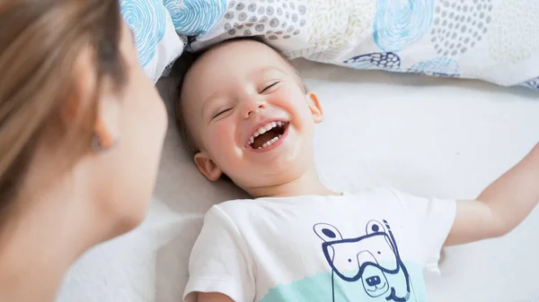 Güzel, genç bir anne ve küçük bir çocuk sabah birlikte eğleniyorlar. Gülümsüyor, gülüyor ve çocukla yatakta oynuyor. Aile mutluluğu konsepti. Parlak, güneşli sıcak bir gün..