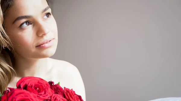 Kırmızı gül buketi olan güzel bir kadın. Şehvetli ve çekici bir kadın gülümseyerek evde çiçeklerin tadını çıkarıyor. Romantik ve kadınsı bir kavram. Yakışıklı portre fotoğrafı..