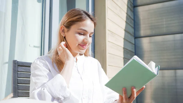 Güneşli bir günde balkonunda kitap okuyan çekici genç bir kadın. Beyaz gömlek giyen ve elinde yeşil bir kitap tutan güzel bir kız..
