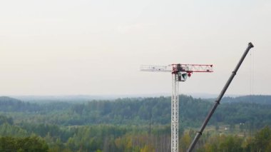Bir kamyon vincinin ve işçilerin yeşil bir vadideki inşaat sahasında orman ve gökyüzünün arka planına karşı kırmızı-beyaz bir vinç monte etmelerinin havadan ve panoramik zamanlaması.