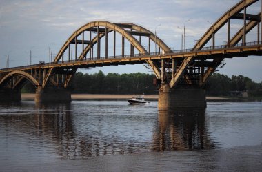 A small boat sails under the bridge of a small river. Small town on river bank
