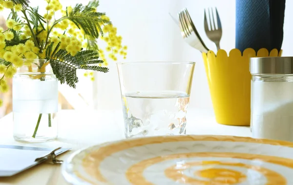 Casual wooden restaurant table with cutlery, glass, yellow flowers, menu and salt shaker