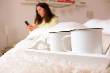Yatakta cep telefonuna bakan bir kadın ve iki beyaz fincan kahve.