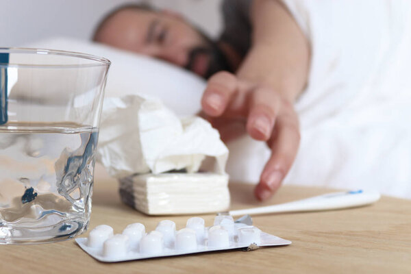 Больной человек в постели с белыми простынями. Над столом таблетки, носовые платки, термометр и стакан воды.
