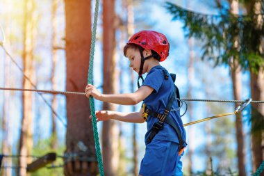 Orman macera parkında bir çocuk. Kırmızı kasklı ve mavi tişörtlü çocuk yüksek ip izine tırmanıyor. Çeviklik becerileri ve çocuklar için açık hava eğlence merkezine tırmanma. Genç çocuk dışarıda oynuyor..
