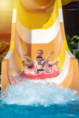 European boy and mom gliding down slide in waterpark. They enjoy the fun and holding hands wide open. clipart