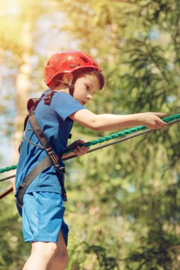 Çocuk güneşli yaz gününde macera parkında eğleniyor. Çocuk halatla oyun sahasına tırmanıyor. Kask sigortalı güvenli tırmanış. Orman macera parkındaki çocuk, ekstrem spor.