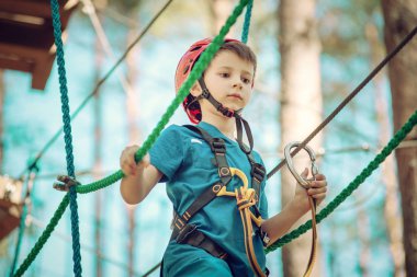 Çocuk güneşli yaz gününde macera parkında eğleniyor. Çocuk halatla oyun sahasına tırmanıyor. Kask sigortalı güvenli tırmanış. Orman macera parkındaki çocuk, ekstrem spor.