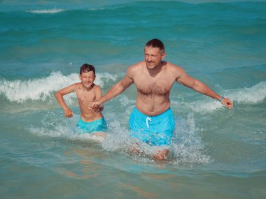 Baba ve oğul aynı şortla güzel deniz kıyısında yürüyorlar. El ele tutuşup mükemmel tatillerinin tadını çıkarıyorlar..