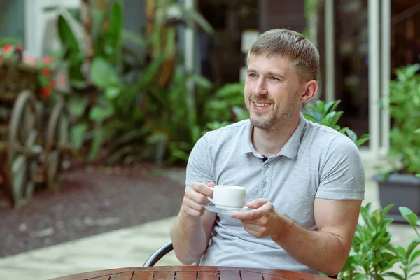 Genç başarılı pozitif adam açık havada bir restoranda oturuyor, kahve içiyor ve hafta sonunun tadını çıkarıyor..