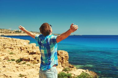 Adam denize bakıyor. Resimli kayalık sahilde ve kollarını sonuna kadar açık tutuyor..