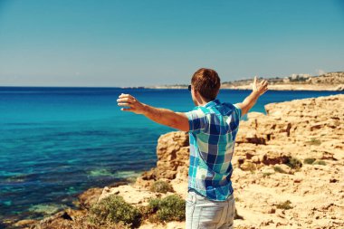 Adam denize bakıyor. Resimli kayalık sahilde ve kollarını sonuna kadar açık tutuyor..