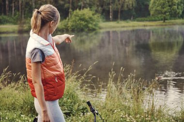 Bisikletinin yanındaki genç bayan bisikletçi parktaki göle bakıyor ve üzerinde bir şey gösteriyor..