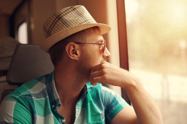 Güneş şapkalı ve güneş gözlüklü bir adam trenle seyahat ediyor. Pencereye bakıyor ve bir şeyler düşünüyor..