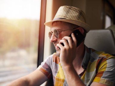 Güneş şapkalı, kareli gömlekli ve güneş gözlüklü yakışıklı trenle seyahat ederken telefonla konuşuyor..