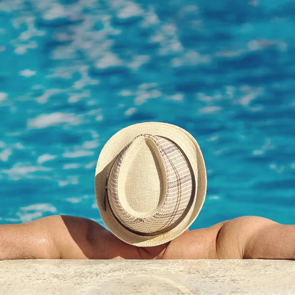 Man in sun hat resting on the side of the swimming pool.