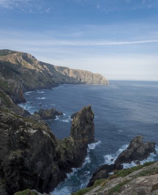 Kıyı manzarası bulutlu, yumuşak dalgalar alçak bitki örtüsüyle kayalıklarda kırılıyor.