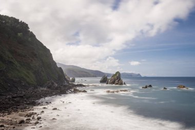 Gökyüzü bulutlu kıyı manzarası, çeşitli adacıklar, yumuşak dalgalar alçak bitki örtüsüyle kayalıklarda kırılır, ipek etkisi
