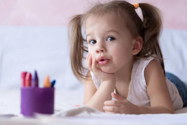 Schöne Mädchen zeichnet mit Bleistiften im Bett. Kleines Mädchen von zwei Jahren malt Malbuch auf dem Bett liegend. Kleines Baby mit Pferdeschwanz, lächelnd auf dem Bett liegend. Hübsches Mädchen malt mit Bleistiften und — Stockfoto